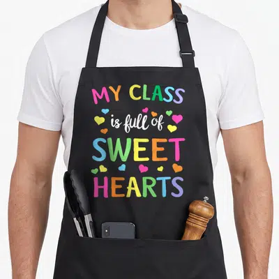 Teacher valentine's day apron - Stacked colorful text MY CLASS is full of SWEET HEARTS with scattered pink orange — shown on a black cotton canvas apron against a white background