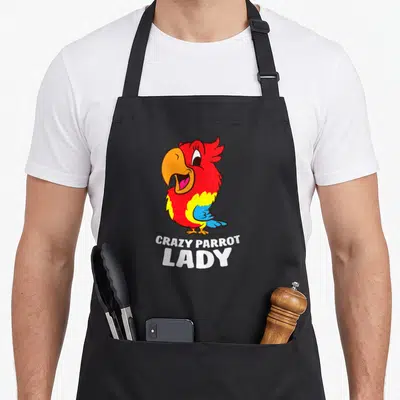 Obsessed parrot lady apron - Cartoon red parrot with yellow belly and blue wing above white text reading CRAZY PARROT — shown on a black cotton canvas apron against a white background