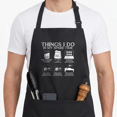 Spare bread apron - White line art showing feeding sourdough, buying flour, and dreaming about bread recipes — shown on a black cotton canvas apron against a white background