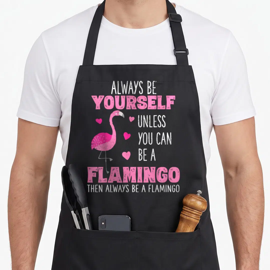 Funny animal flamingo apron - Pink cartoon flamingo with four hearts and text Always Be Yourself Unless You Can Be — shown on a black cotton canvas apron against a white background