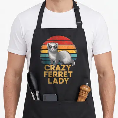 Sunglass crazy ferret man apron - Grey ferret wearing sunglasses stands before retro striped sun with stacked yellow — shown on a black cotton canvas apron against a white background