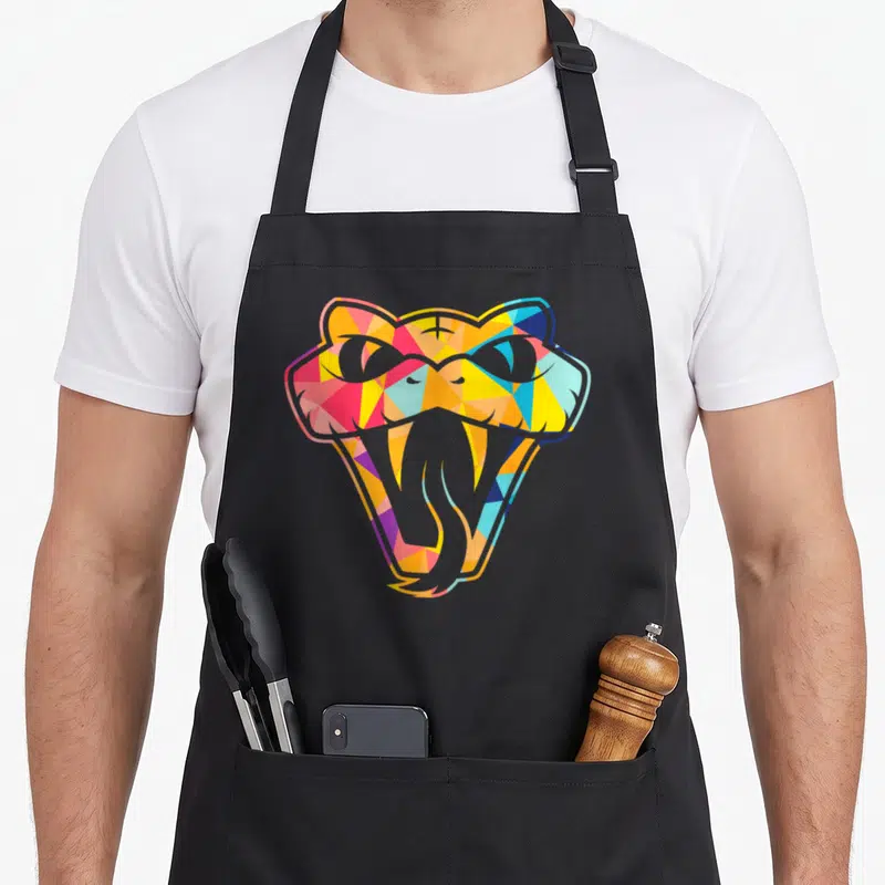 Snake colourful apron - Stylized snake head with open mouth filled with yellow, pink, blue, orange, and grey geometric — shown on a black cotton canvas apron against a white background