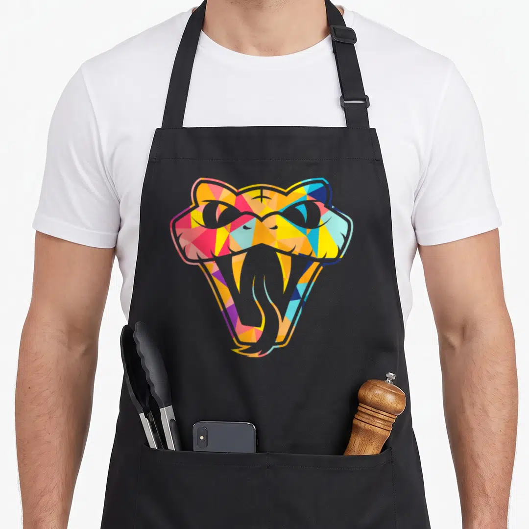 Snake colourful apron - Stylized snake head with open mouth filled with yellow, pink, blue, orange, and grey geometric — shown on a black cotton canvas apron against a white background