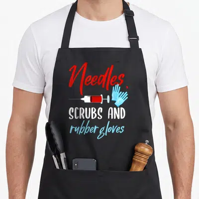 Phlebotomist gloves and apron - Needles, scrub and rubber gloves text with cartoon syringe and blue gloves — shown on a black cotton canvas apron against a white background