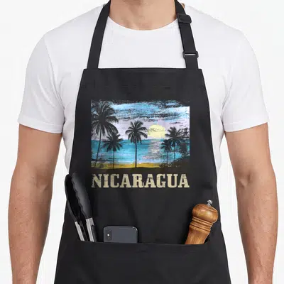 Surfer nicaragua apron - Retro beach scene with palm trees and NICARAGUA text in blue beige grey yellow — shown on a black cotton canvas apron against a white background