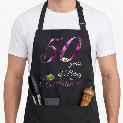 50th floral aprons - Purple 50 with daisy bee, stacked white text, and yellow rose overlapping the word Awesome — shown on a black cotton canvas apron against a white background