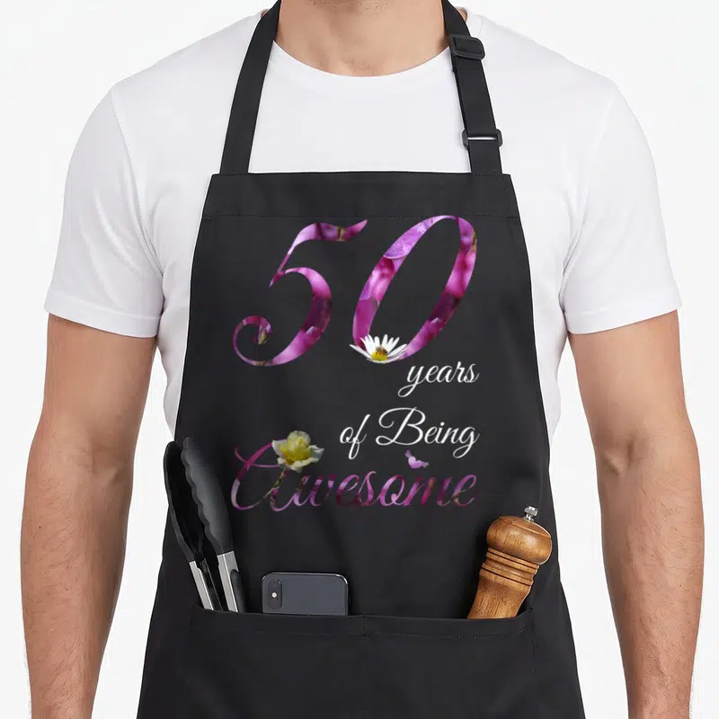 50th floral aprons - Purple 50 with daisy bee, stacked white text, and yellow rose overlapping the word Awesome — shown on a black cotton canvas apron against a white background