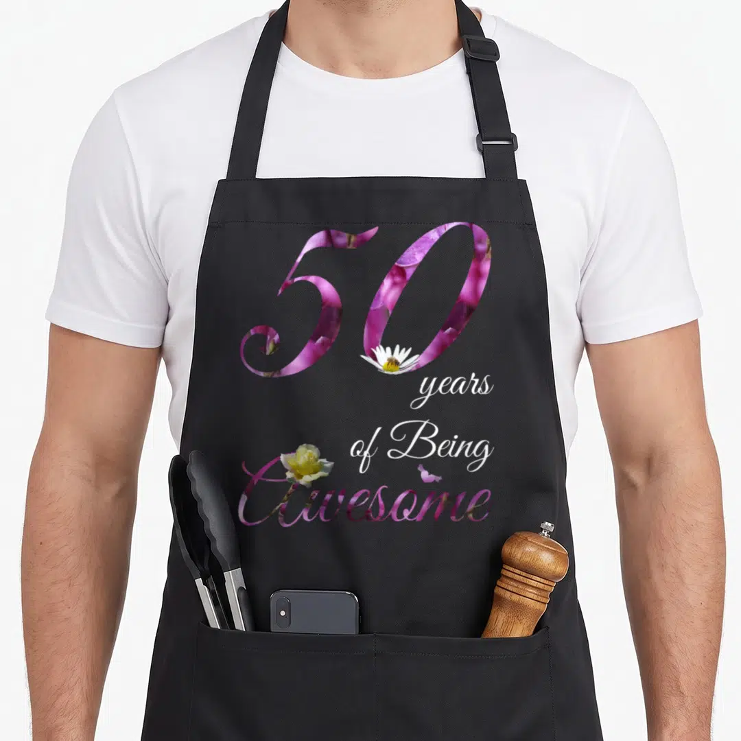50th floral aprons - Purple 50 with daisy bee, stacked white text, and yellow rose overlapping the word Awesome — shown on a black cotton canvas apron against a white background