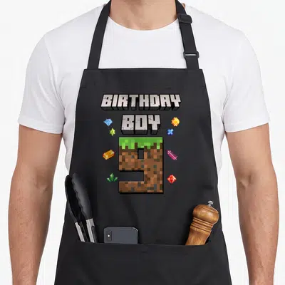 9th video birthday apron - Pixelated BIRTHDAY BOY text and dirt-textured number 9 surrounded by colorful gems — shown on a black cotton canvas apron against a white background