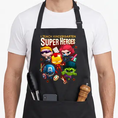 Shot heroe apron - Four chibi superheroes surrounded by pencils, scissors, and an A+ paper test — shown on a black cotton canvas apron against a white background