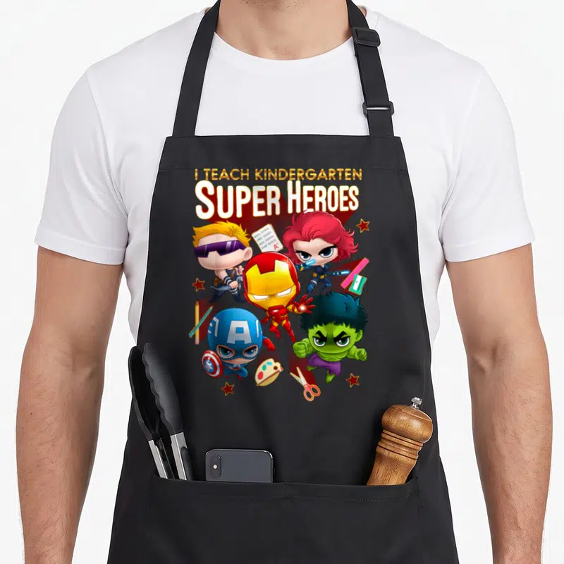 Shot heroe apron - Four chibi superheroes surrounded by pencils, scissors, and an A+ paper test — shown on a black cotton canvas apron against a white background