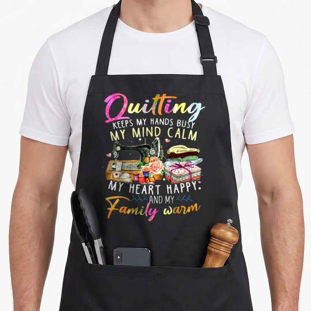 Quilting busy apron - Vintage sewing machine with thread spools and fabric stack surrounded by pink and yellow text — shown on a black cotton canvas apron against a white background