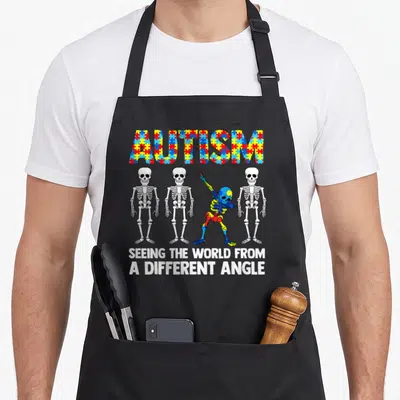 Funny awareness autism apron - Colored puzzle piece AUTISM text above three white skeletons and one leaning — shown on a black cotton canvas apron against a white background
