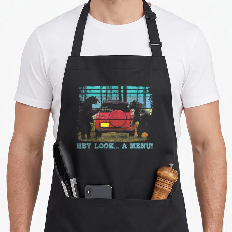 Hey happy camper apron - Two bears facing a red car with distressed text HEY LOOK... A MENU! — shown on a black cotton canvas apron against a white background