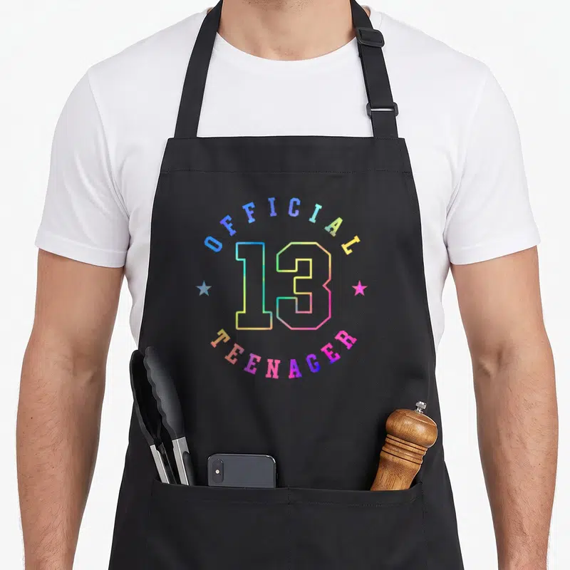 Thirteen colorful aprons - Rainbow number 13 with Official Teenager text and two flanking stars — shown on a black cotton canvas apron against a white background