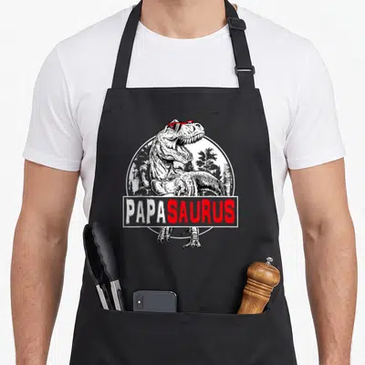 Papasaurus t rex apron - Line-art T-Rex with red sunglasses and PAPASAURUS text over a forest circle — shown on a black cotton canvas apron against a white background