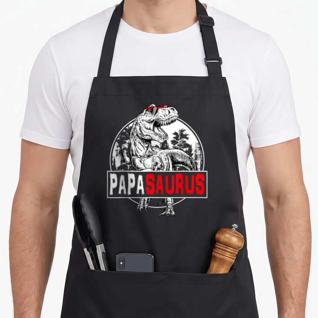 Papasaurus t rex apron - Line-art T-Rex with red sunglasses and PAPASAURUS text over a forest circle — shown on a black cotton canvas apron against a white background