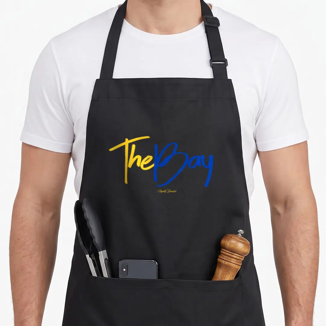 Bay curry apron - Yellow and blue script text The Bay Royally Branded stacked vertically — shown on a black cotton canvas apron against a white background