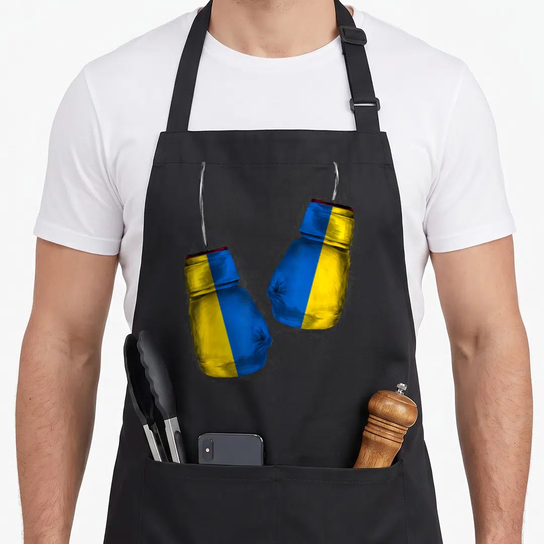 Boxer ukrainian aprons - Two boxing gloves in blue yellow and red hanging from silver hooks with black splatters — shown on a black cotton canvas apron against a white background