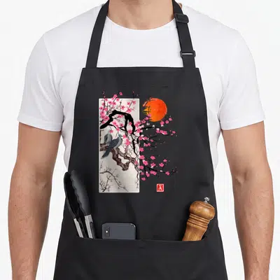 Scenery cherry apron - Two grey birds on black branch with pink flowers, orange sun, and red seal inside white frame — shown on a black cotton canvas apron against a white background