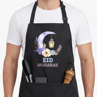 Mubarak eid apron - Watercolor crescent moon, hanging lantern, purple and yellow flowers, and stacked EID MUBARAK text — shown on a black cotton canvas apron against a white background