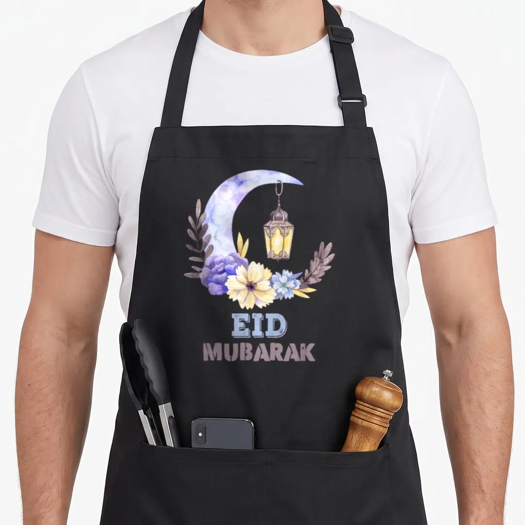 Mubarak eid apron - Watercolor crescent moon, hanging lantern, purple and yellow flowers, and stacked EID MUBARAK text — shown on a black cotton canvas apron against a white background