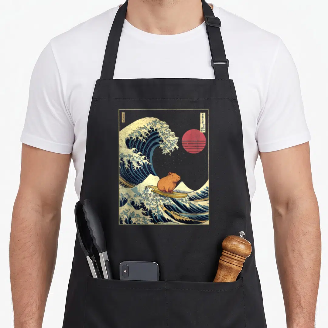 Japanese animal apron - Capybara surfing blue wave with red sun and vertical Japanese text in retro style — shown on a black cotton canvas apron against a white background