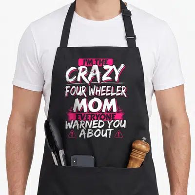 Four mother's apron - Pink and white typography reading I'M THE CRAZY FOUR WHEELER MOM with warning triangles — shown on a black cotton canvas apron against a white background