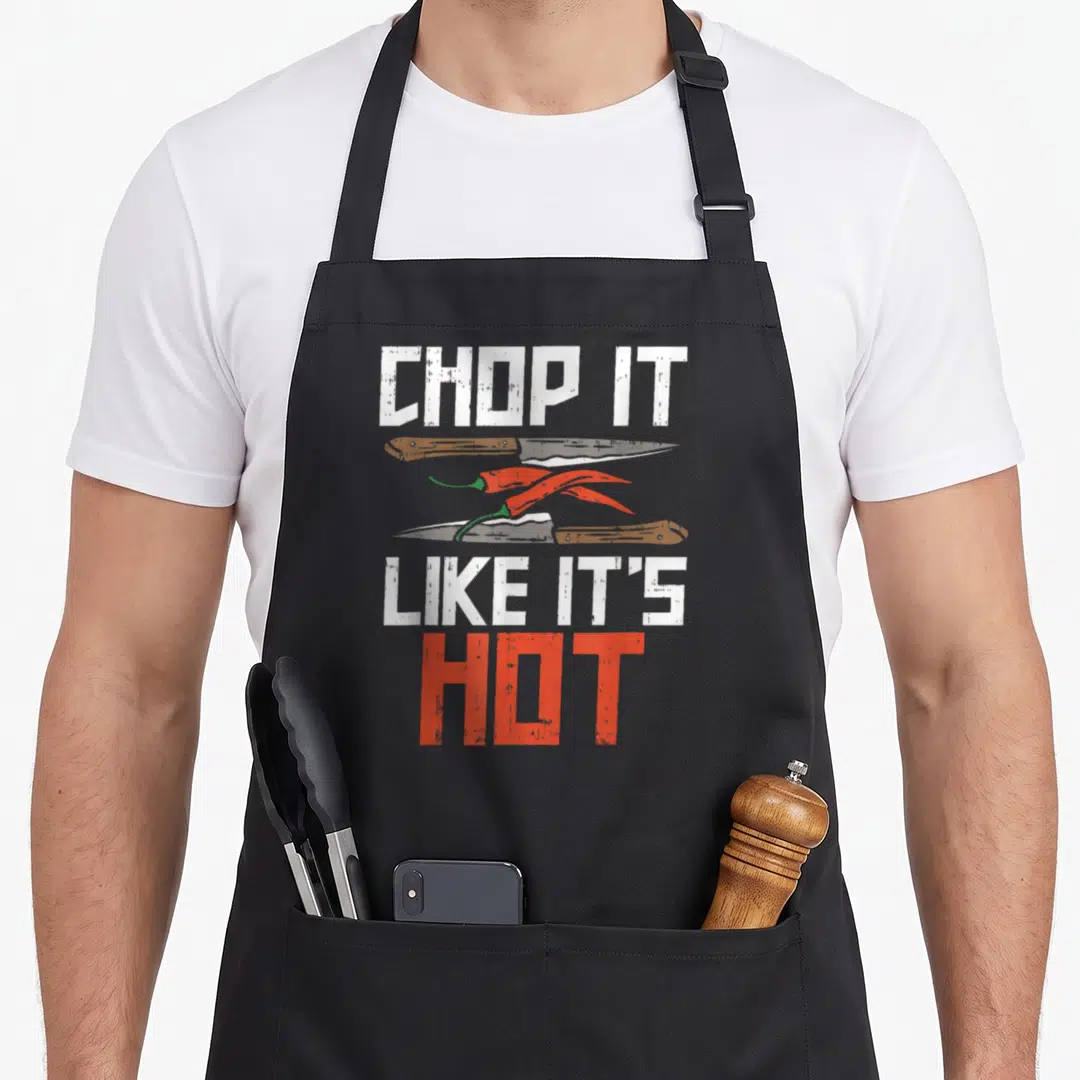 Funny camping culinary aprons - CHOP IT LIKE IT'S HOT text flanking two knives and red chili peppers in distressed print — shown on a black cotton canvas apron against a white background