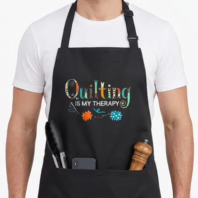 Quilting my therapy apron - Patchwork text reading Quilting IS MY THERAPY above two pincushions and a needle — shown on a black cotton canvas apron against a white background