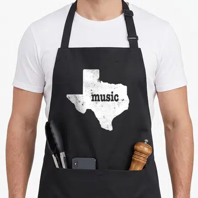 Country texas aprons - Distressed white Texas map silhouette with grey music text centered inside — shown on a black cotton canvas apron against a white background