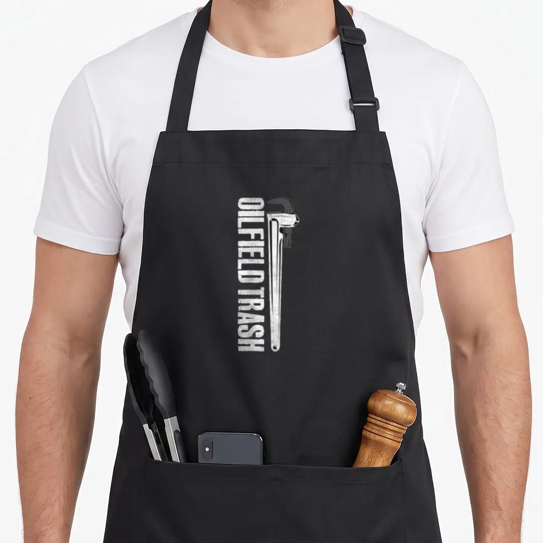 Oilfield field life apron - Vertical white OILFIELD TRASH text beside a distressed pipe wrench illustration — shown on a black cotton canvas apron against a white background