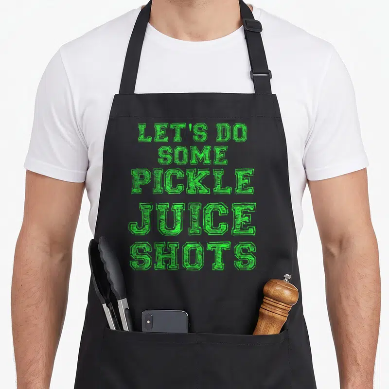 Juice pickle apron - Green stacked text reading LET'S DO SOME PICKLE JUICE SHOTS in block letters — shown on a black cotton canvas apron against a white background