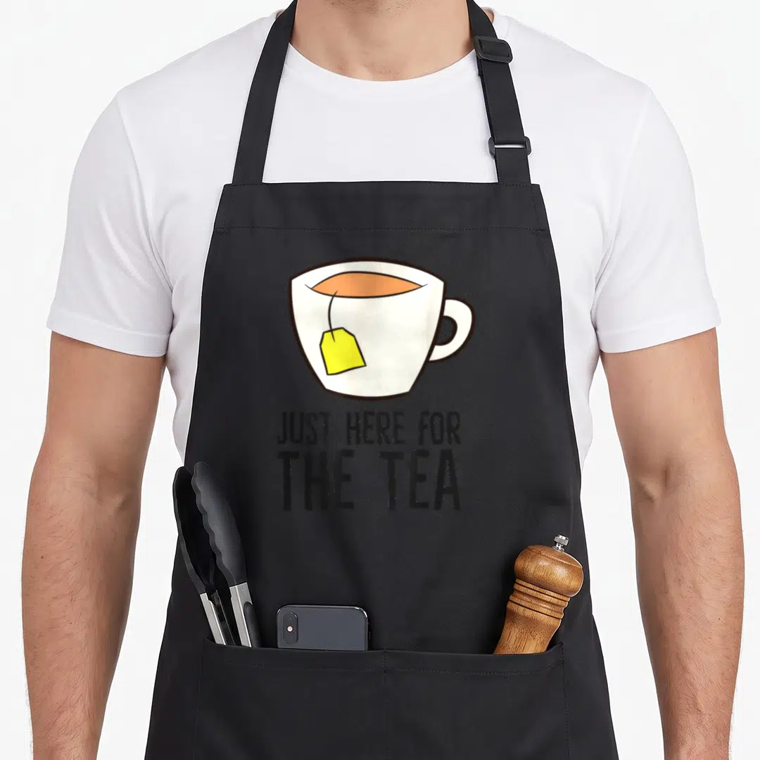 Funny drinking tea party aprons - Cartoon white cup with yellow tea tag and black text reading JUST HERE FOR THE TEA — shown on a black cotton canvas apron against a white background