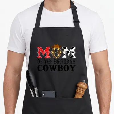 1st birthday cowboy apron - MOM OF THE BIRTHDAY COWBOY text with cowboy hat, lasso, paisley, and cow print patterns — shown on a black cotton canvas apron against a white background