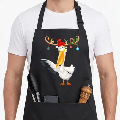 Pelican reindeer christmas apron - Cartoon pelican with antlers, ornaments, lights, and Santa hat — shown on a black cotton canvas apron against a white background