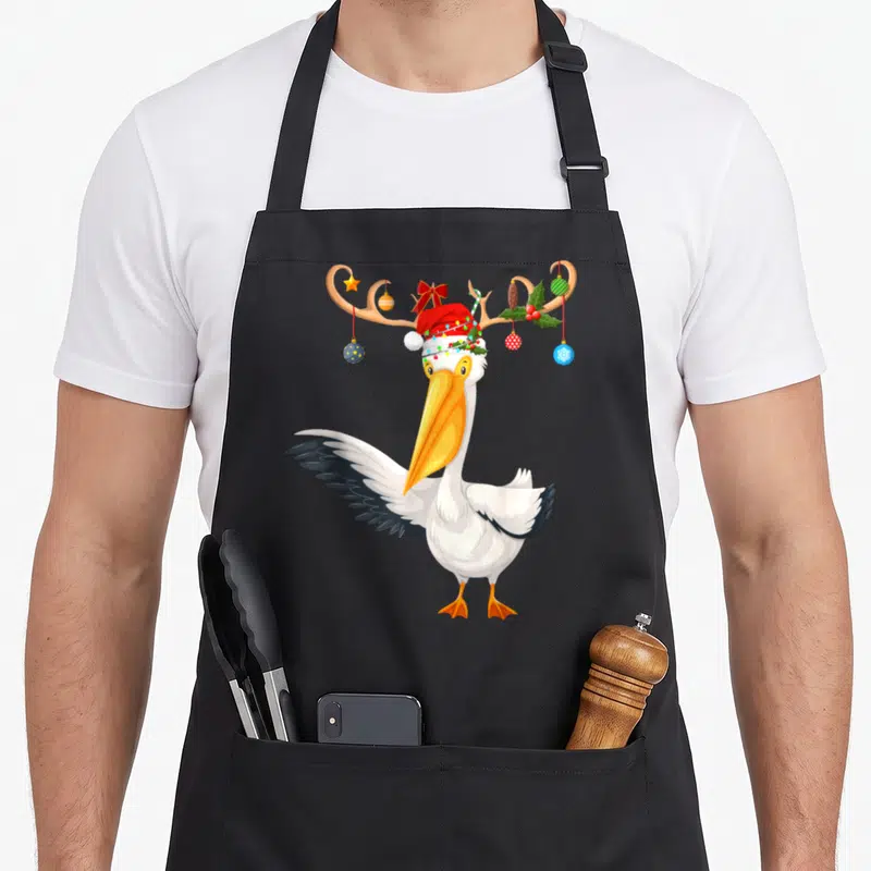 Pelican reindeer christmas apron - Cartoon pelican with antlers, ornaments, lights, and Santa hat — shown on a black cotton canvas apron against a white background