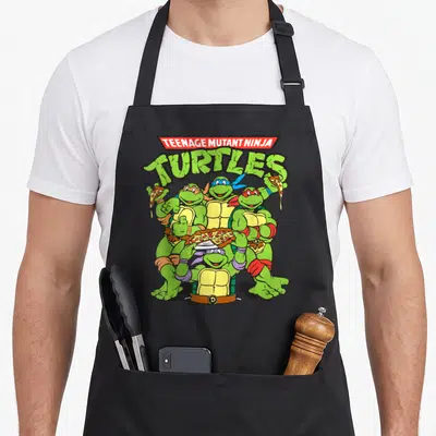 Cute pizza apron - Stacked TMNT text above four turtles holding pizza slices in green red and yellow — shown on a black cotton canvas apron against a white background
