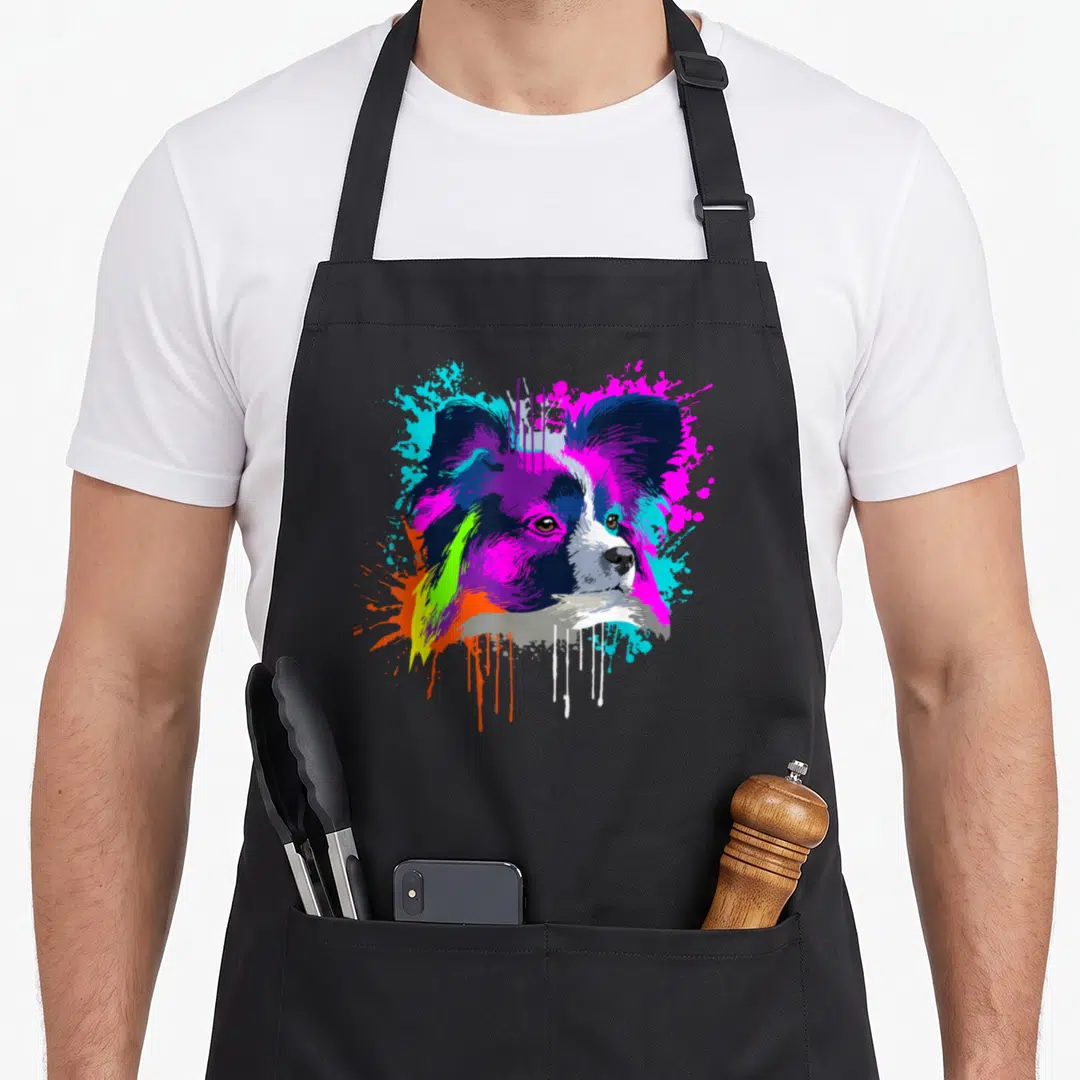 Splash papillon dog apron - Cartoon papillon head with purple magenta cyan orange and white paint splatters — shown on a black cotton canvas apron against a white background
