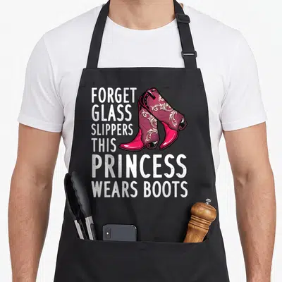 Rodeo western apron - White text reading Forget Glass Slippers This Princess Wears Boots next to two magenta cowboy — shown on a black cotton canvas apron against a white background
