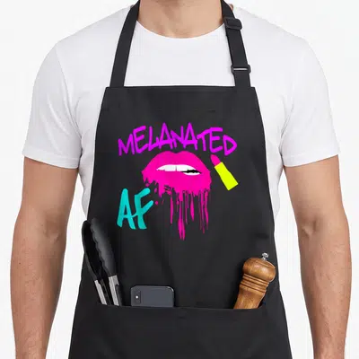 Melanated drip apron - MELANATED AF text over magenta lips with white teeth, cyan letters, yellow lipstick, cartoon — shown on a black cotton canvas apron against a white background