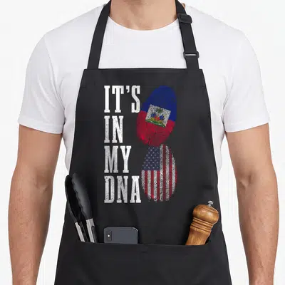 Haiti dna american flag apron - Stacked fingerprints filled with Haitian and USA flags beside vertical IT'S IN MY DNA — shown on a black cotton canvas apron against a white background