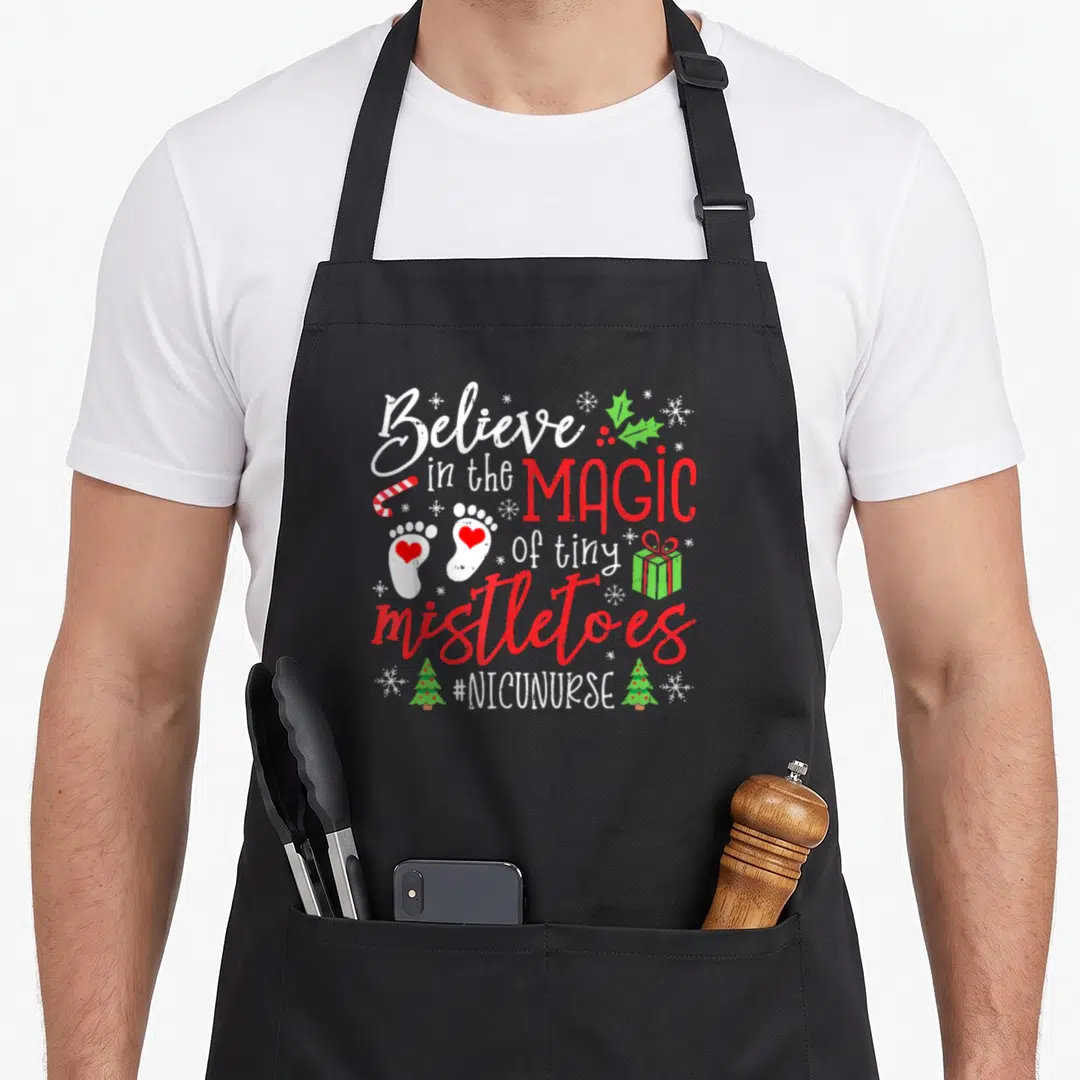 Nicu christmas apron - Believin Magic of Tiny Mistletoes text with baby footprints, candy cane, and holly — shown on a black cotton canvas apron against a white background