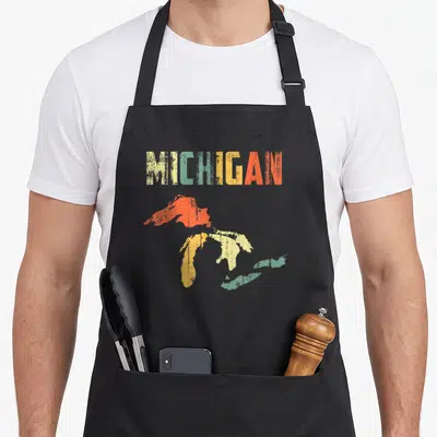 Lake pride apron - MICHIGAN text above yellow green orange and red distressed state map silhouette — shown on a black cotton canvas apron against a white background
