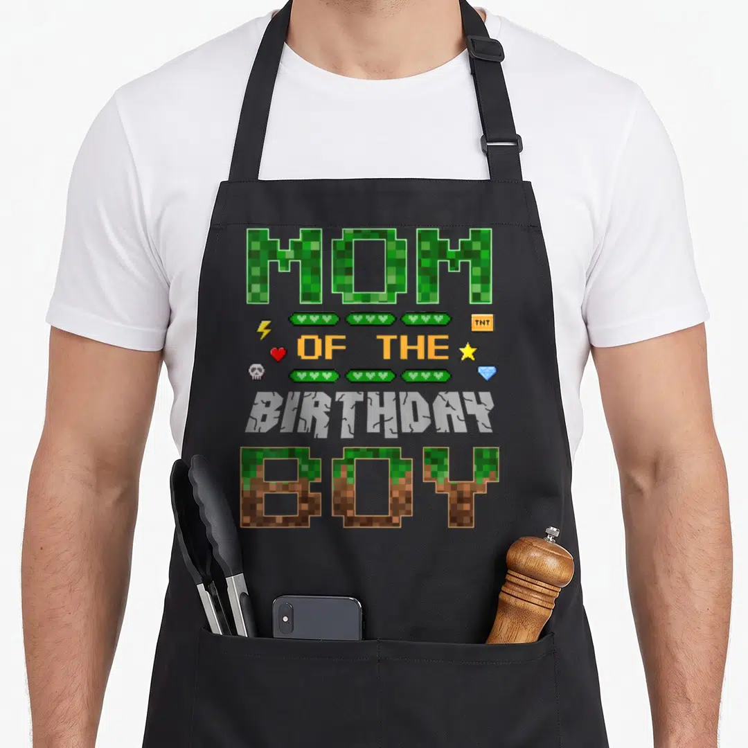 Pixel family aprons - Pixel text MOM OF THE BIRTHDAY BOY with health bars and game icons in green brown orange grey — shown on a black cotton canvas apron against a white background