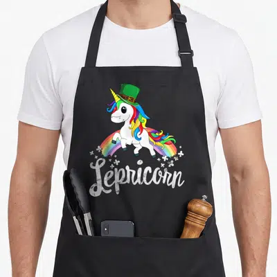 Unicorn st patrick day aprons - Cartoon white unicorn with rainbow mane wearing green top hat above rainbow — shown on a black cotton canvas apron against a white background