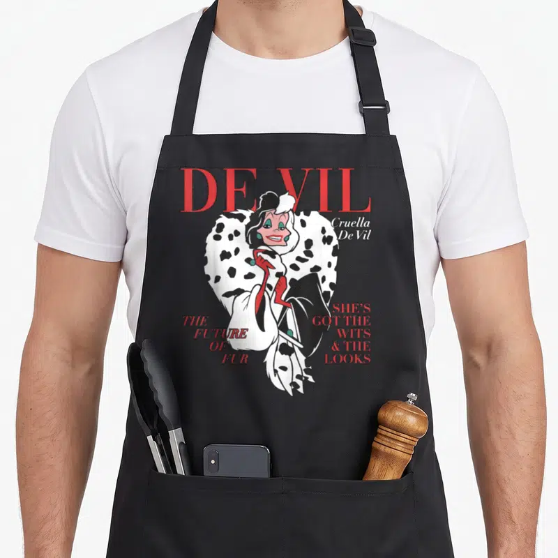 Cruella novelty apron - Cruella cartoon in spotted coat with red DE VIL text and fur fashion quotes — shown on a black cotton canvas apron against a white background