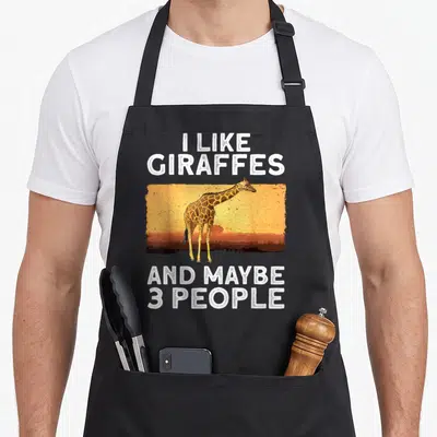 Zoo animal giraffe apron - Cartoon giraffe standing in orange sunset with white text reading I Like Giraffes And Maybe — shown on a black cotton canvas apron against a white background