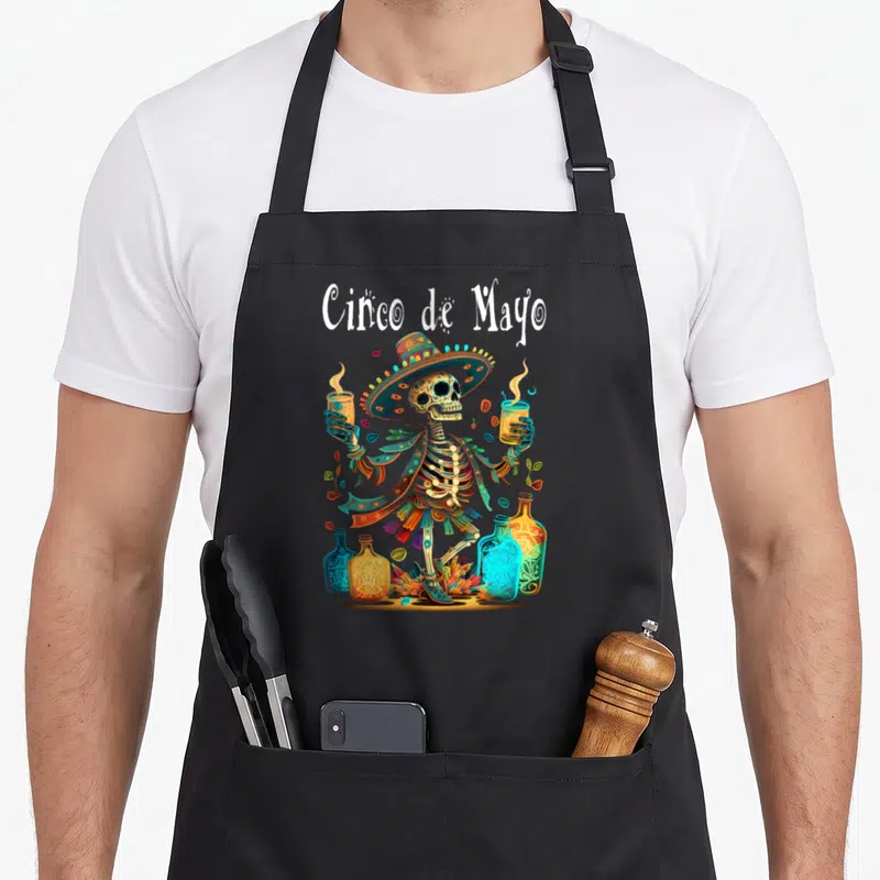 Festival cinco de mayo apron - Cartoon skeleton in sombrero holding two glasses surrounded by bottles and leaves — shown on a black cotton canvas apron against a white background