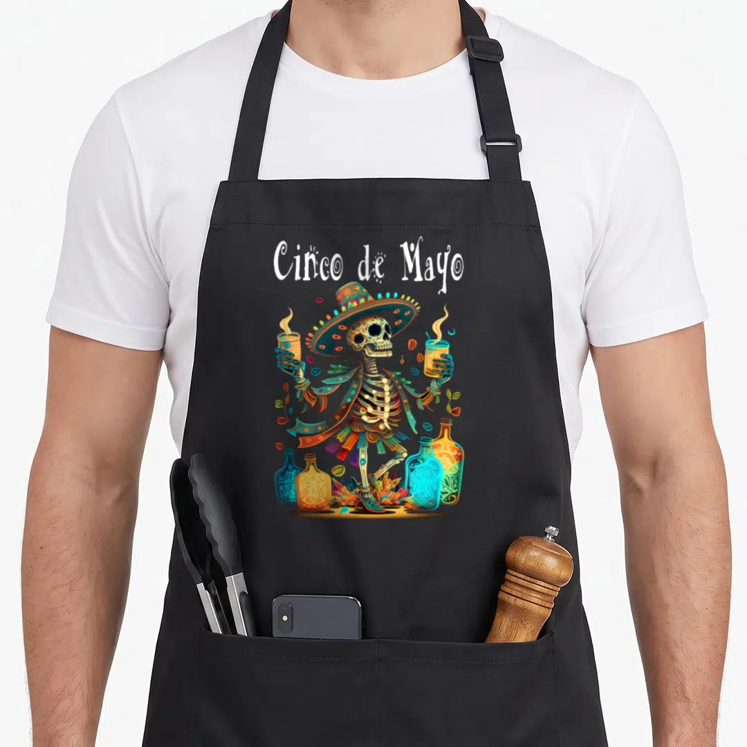 Festival cinco de mayo apron - Cartoon skeleton in sombrero holding two glasses surrounded by bottles and leaves — shown on a black cotton canvas apron against a white background